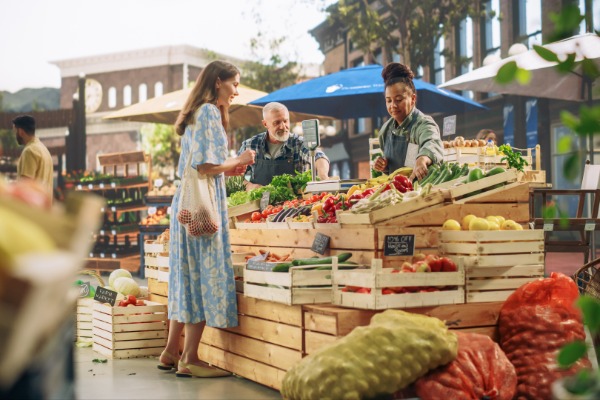 A woman chooses vegetables at an outdoor market stand while two people arrange produce behind wooden crates.