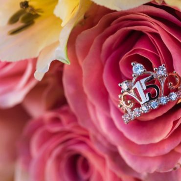 Close-up of a jeweled number 15 tiara nestled in a pink rose among soft pink and pale yellow flowers.