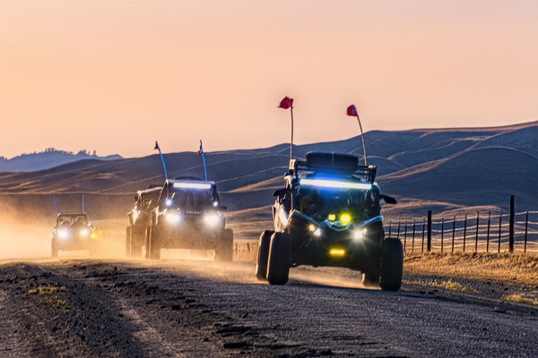 A lineup of four UTVs driving along a dirt road at sunset. Their wheels are kicking up dust clouds behind them.