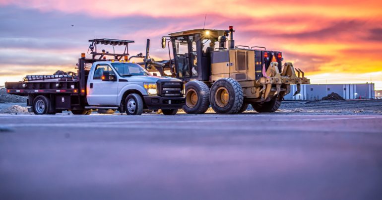 A work truck is parked next to a piece of heavy machinery. The sun is low on the horizon.