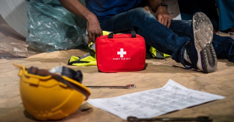 A close-up of a first aid kit on the ground next to a man sitting on a high-visibility vest next to a hard hat and tools.