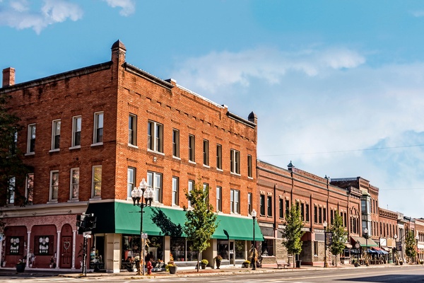 Row of multi-story brick buildings with shops and awnings stretches along a quiet downtown street under a blue sky.