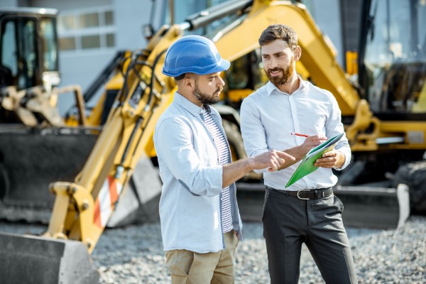 Two men, one in a button-up shirt and the other in a blue hard hat, are standing in the middle of a construction zone.