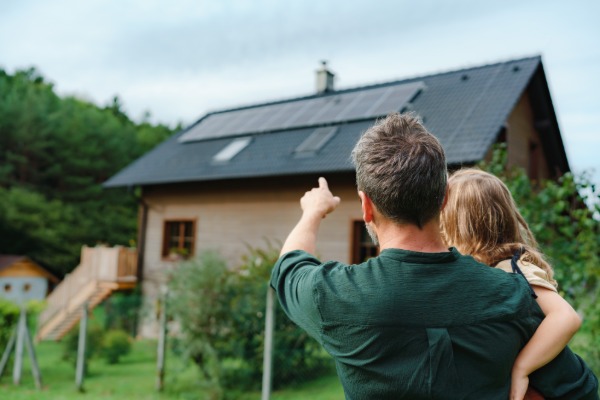 A man holding a small girl, pointing at the top of a house with multiple solar panels surrounded by greenery.