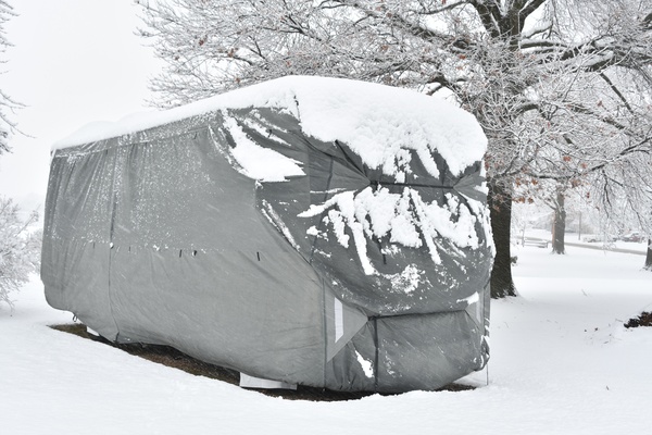 A recreational vehicle camper parked outside with a comprehensive gray cover over it, covered in snow.