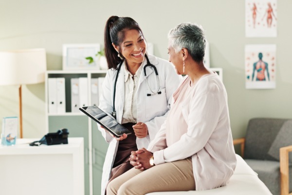 A female doctor wearing a white robe standing next to a senior woman sitting on a small bed inside a room.