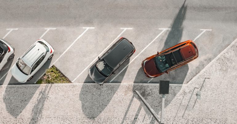 Four cars parked in parking spots along a sidewalk during the daytime. One car is parked across two spots.